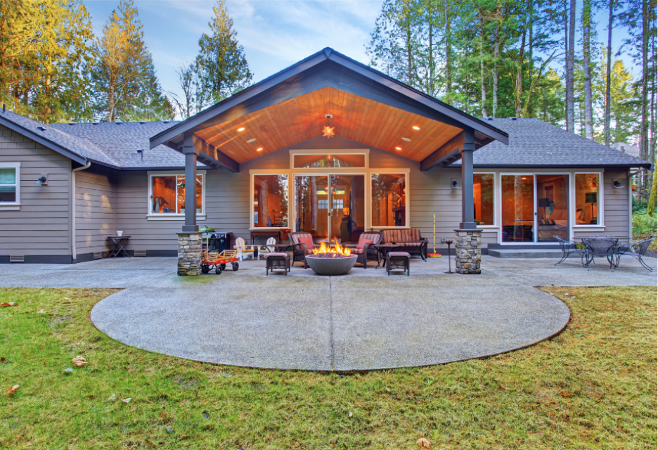 A modern single-story home with a covered outdoor living area featuring comfortable seating, a central fire pit, and a spacious concrete patio surrounded by trees.